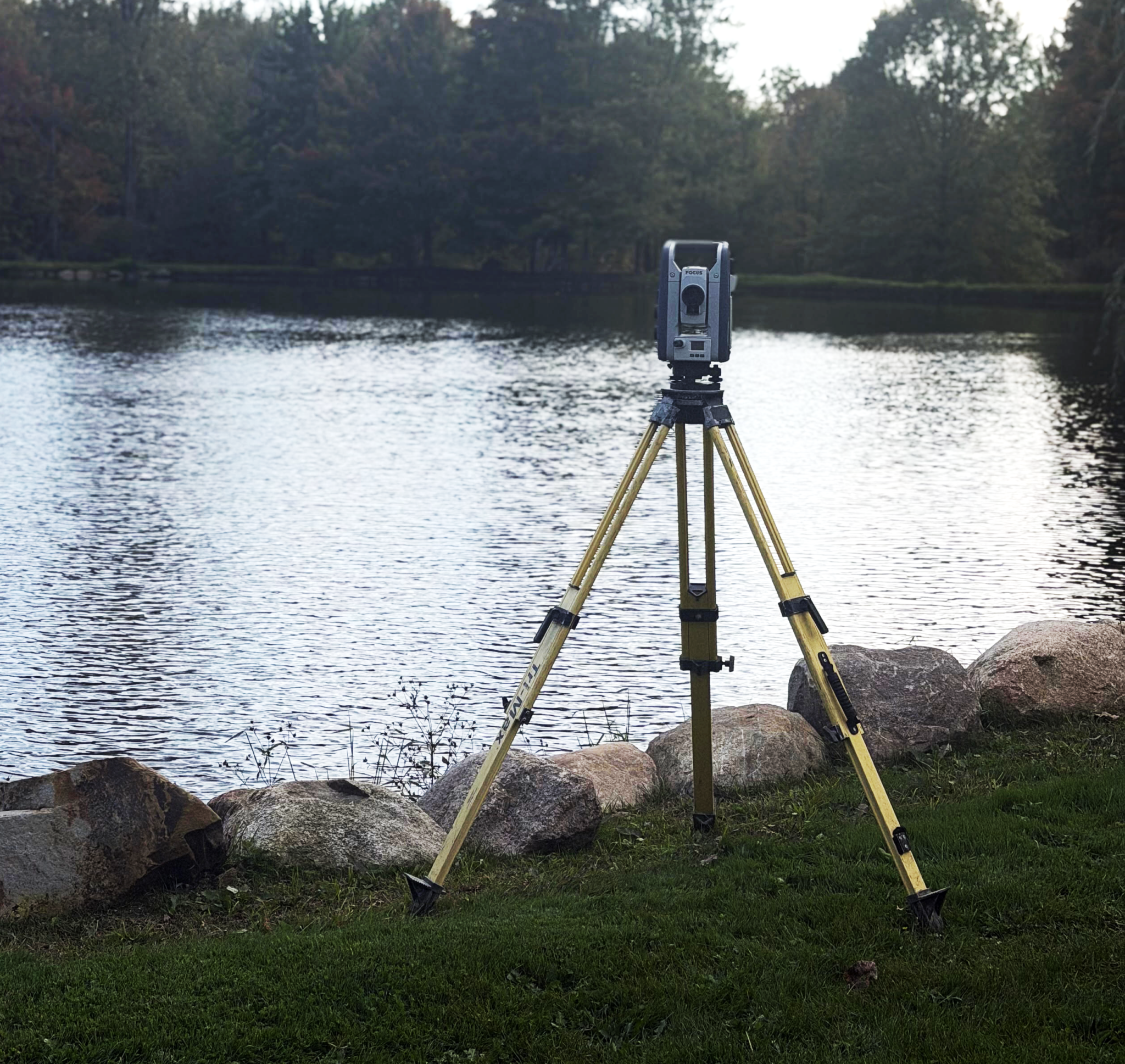 A surveyor uses a tripod-mounted surveying instrument beside a body of water to measure land and distances.