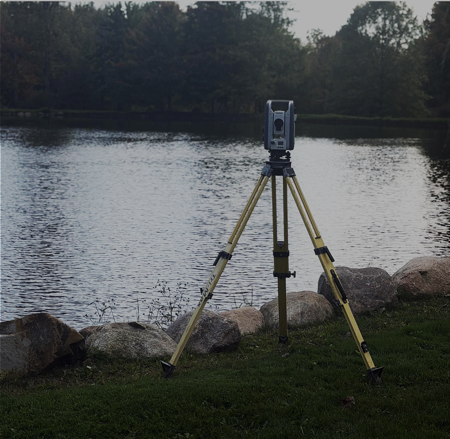 A surveyor uses a tripod-mounted surveying instrument beside a body of water to measure land and distances.